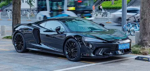 A black McLaren GT parked on a city street beside bicycles, with a bus passing behind it.