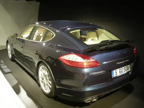 Dark blue Porsche Panamera Turbo lit dramatically on a museum display floor, rear three-quarter view.