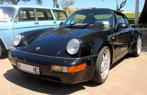 Black Porsche 964 Turbo parked beside a vintage estate car at an outdoor classic car gathering, front three-quarter view.