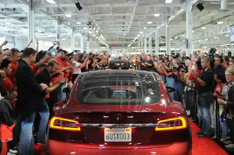 A red Tesla Model S rolling down a red carpet through a cheering crowd at the Fremont factory's first deliveries event.